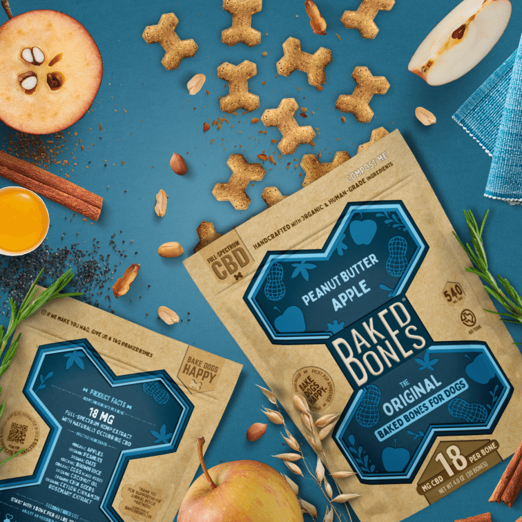 Busy image including golden brown baked dog bones pouring out of two Kraft and blue bags of BakedBones, cut apples, rosemary, shelled peanuts, cinnamon, and chia seeds, all against a blue background