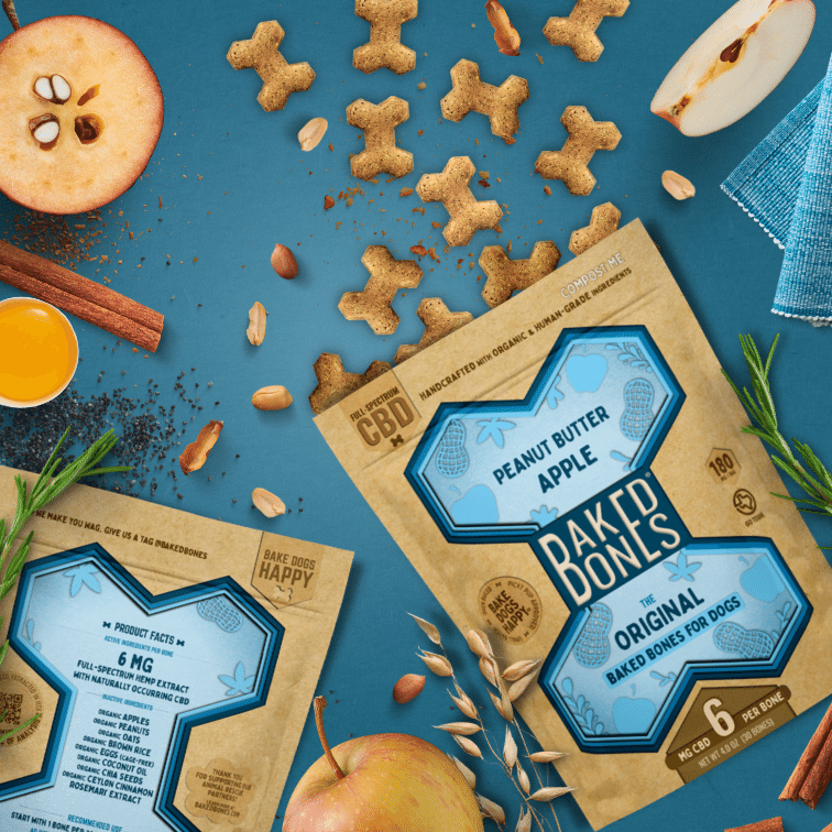Busy image including golden brown baked dog bones pouring out of two Kraft and blue bags of BakedBones, cut apples, rosemary, shelled peanuts, cinnamon, and chia seeds, all against a blue background