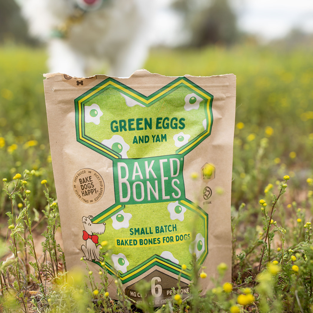 Close up photograph of a kraft BakedBones bag with a large green bone on the front and labeled “Green Eggs and Yam.”  The bag is sitting in a field of green and yellow flowers and there is a dog in the blurry background approaching the bag